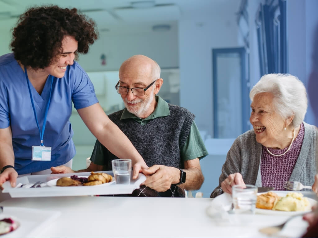 A smiling caregiver in blue scrubs serving a meal tray to an older man and woman seated at a table, with the woman laughing warmly as they share a mealtime together.