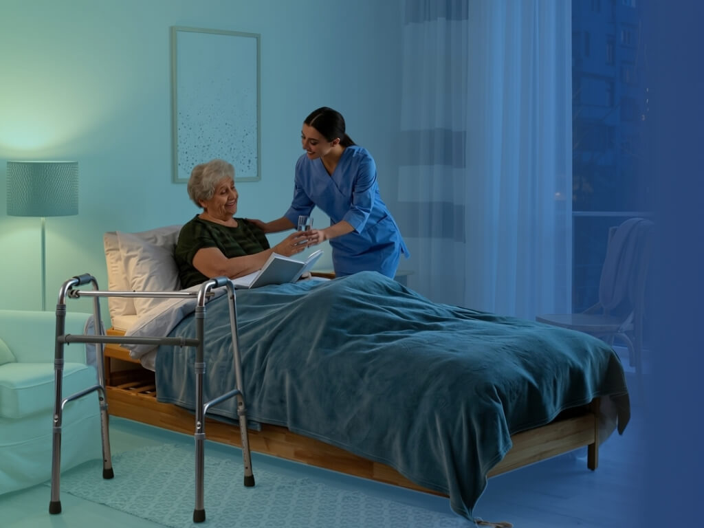 A caregiver in blue scrubs handing a glass of water to a smiling elderly woman resting in bed in the evening, with a walker beside the bed and a lamp glowing in the corner of a calm, softly lit bedroom.