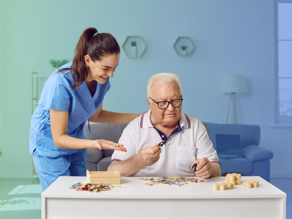 Caregiver in blue scrubs smiling as an older man with glasses works on a wooden puzzle at home during Alzheimer’s care