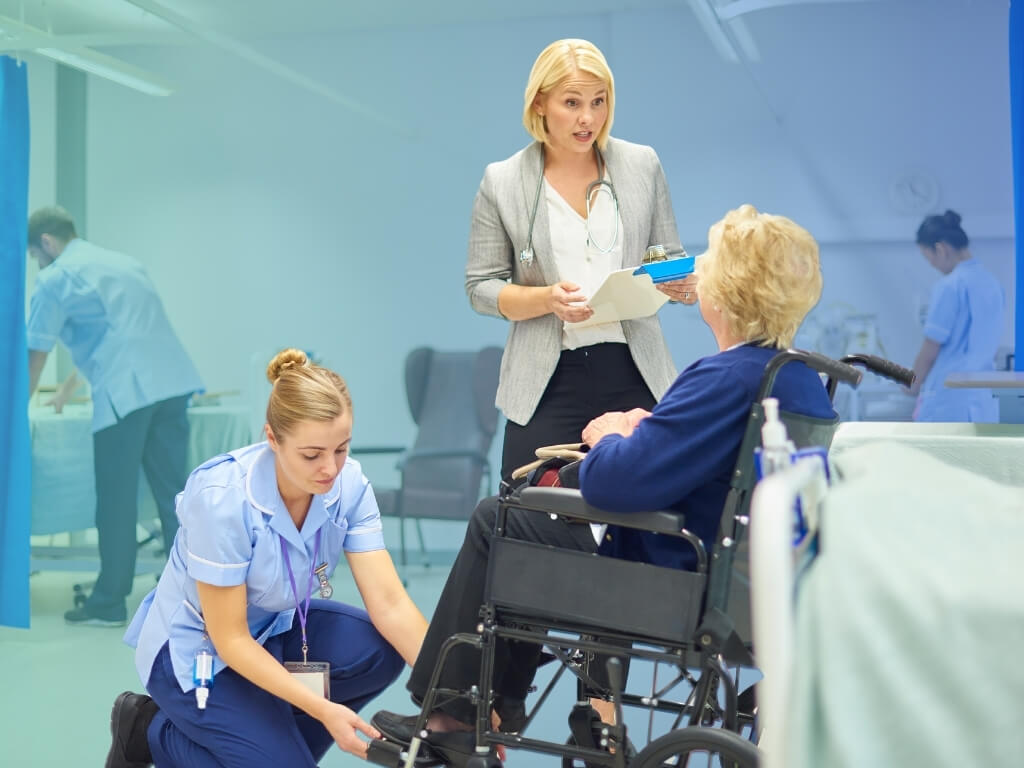 Nurse adjusting a wheelchair footrest for an older woman while a doctor with a clipboard speaks with her in a hospital ward - Post-Hospital Recovery Care