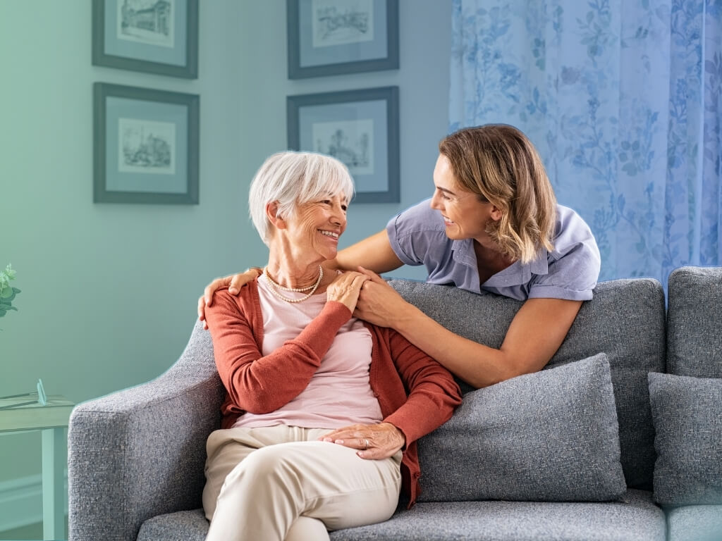 Smiling adult daughter hugging her elderly mother on a sofa at home, a moment of care support for elderly parents