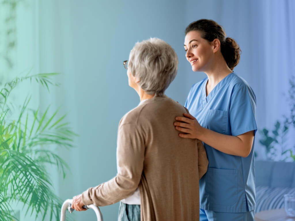 Caregiver encouraging an older woman as she walks with a walker, illustrating mobility assistance and fall prevention
