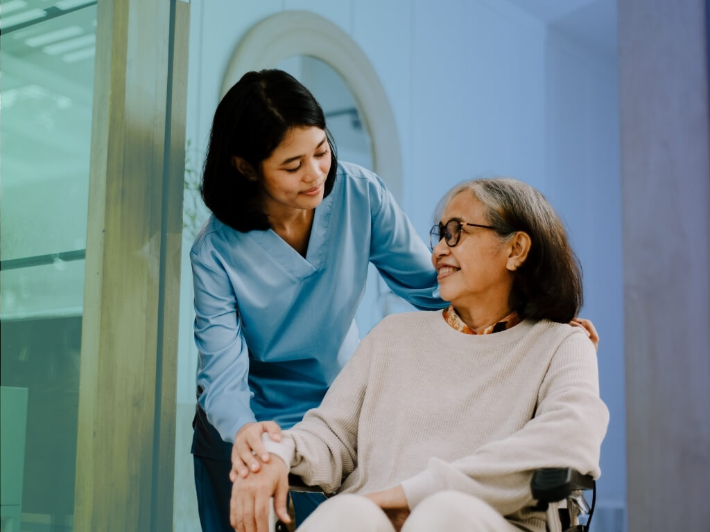 Smiling caregiver beside an older woman in a wheelchair, illustrating respectful incontinence care and mobility support