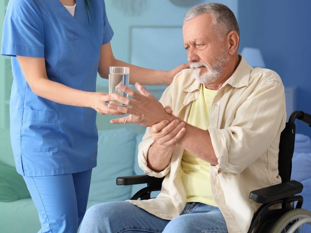 A caregiver in blue scrubs offering a glass of water to an older man in a wheelchair, who steadies his hand with his other hand, in a bright, comfortable home setting.