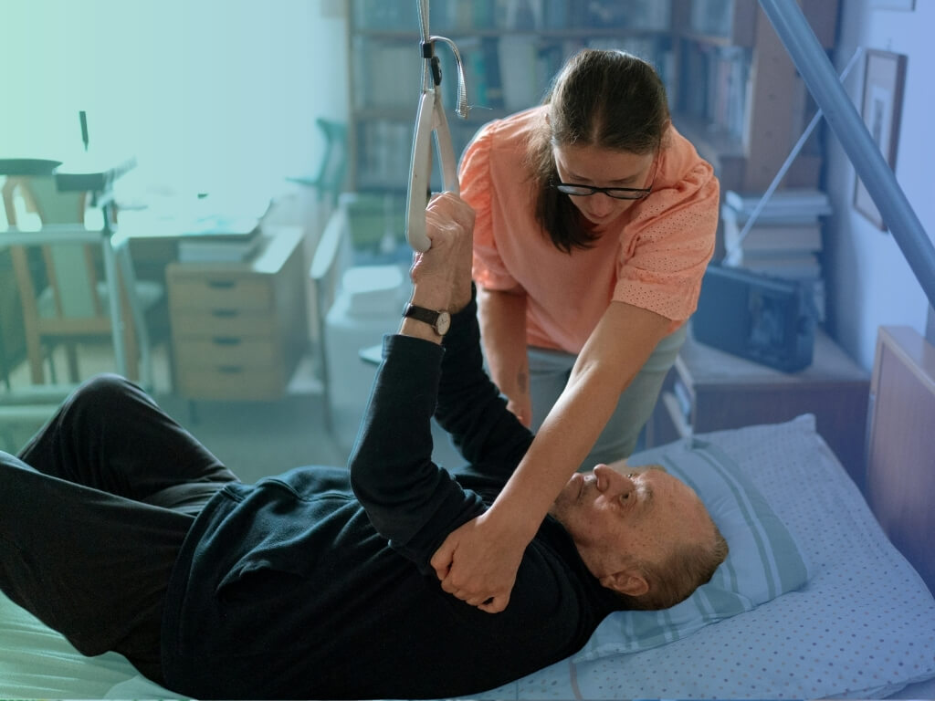 Caregiver helping an older man reposition in bed using an overhead trapeze bar during home recovery - Post-Hospital Recovery Care