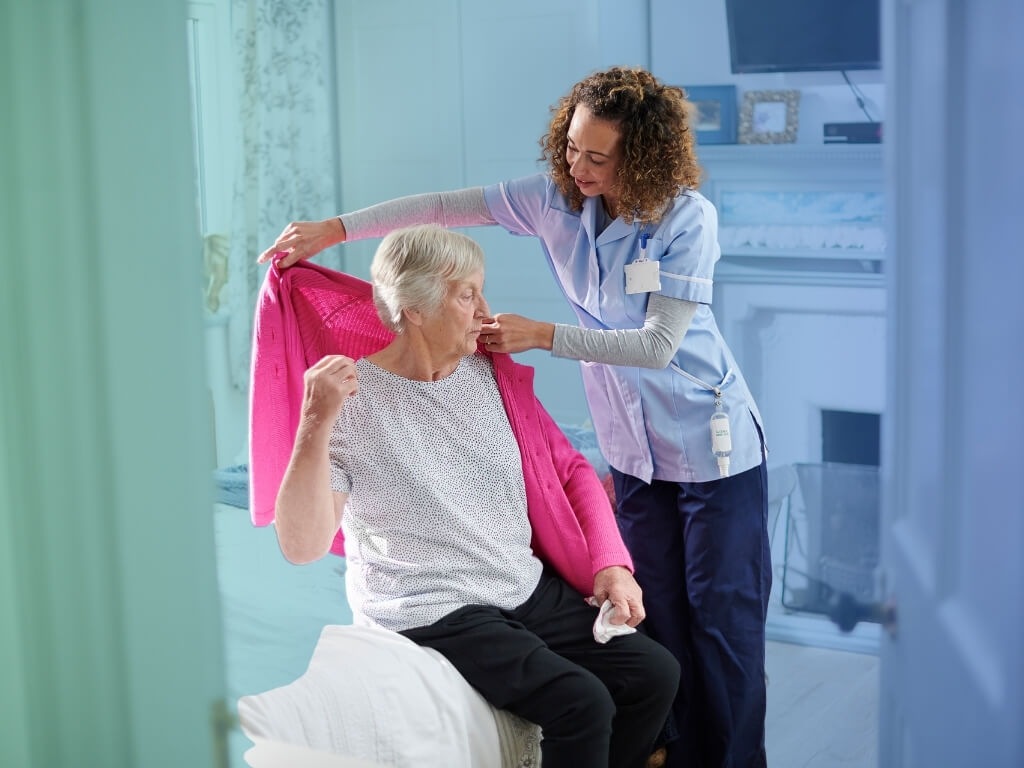 Respite Care -Caregiver helping an older woman put on a pink cardigan while she sits on the edge of her bed at home