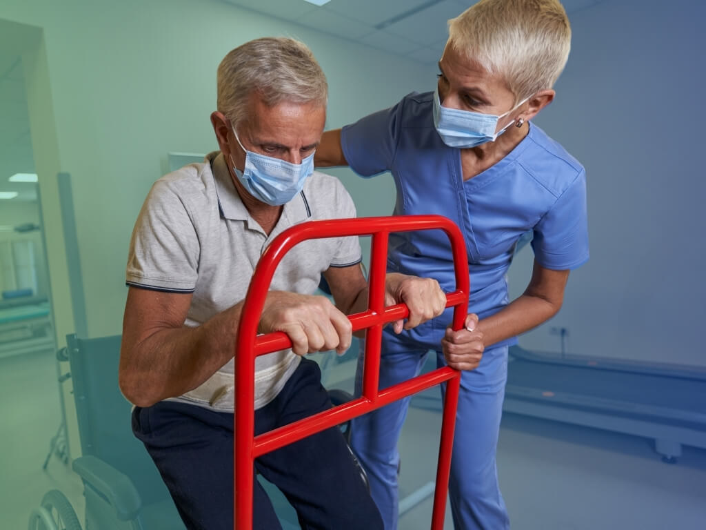 Stroke Recovery Care - Caregiver in a face mask supporting an older man as he stands up using a red mobility support frame