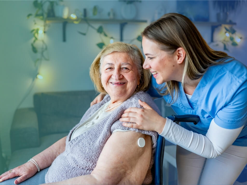 Caregiver in blue scrubs smiling beside an older woman at home, supporting an older adult with a chronic condition